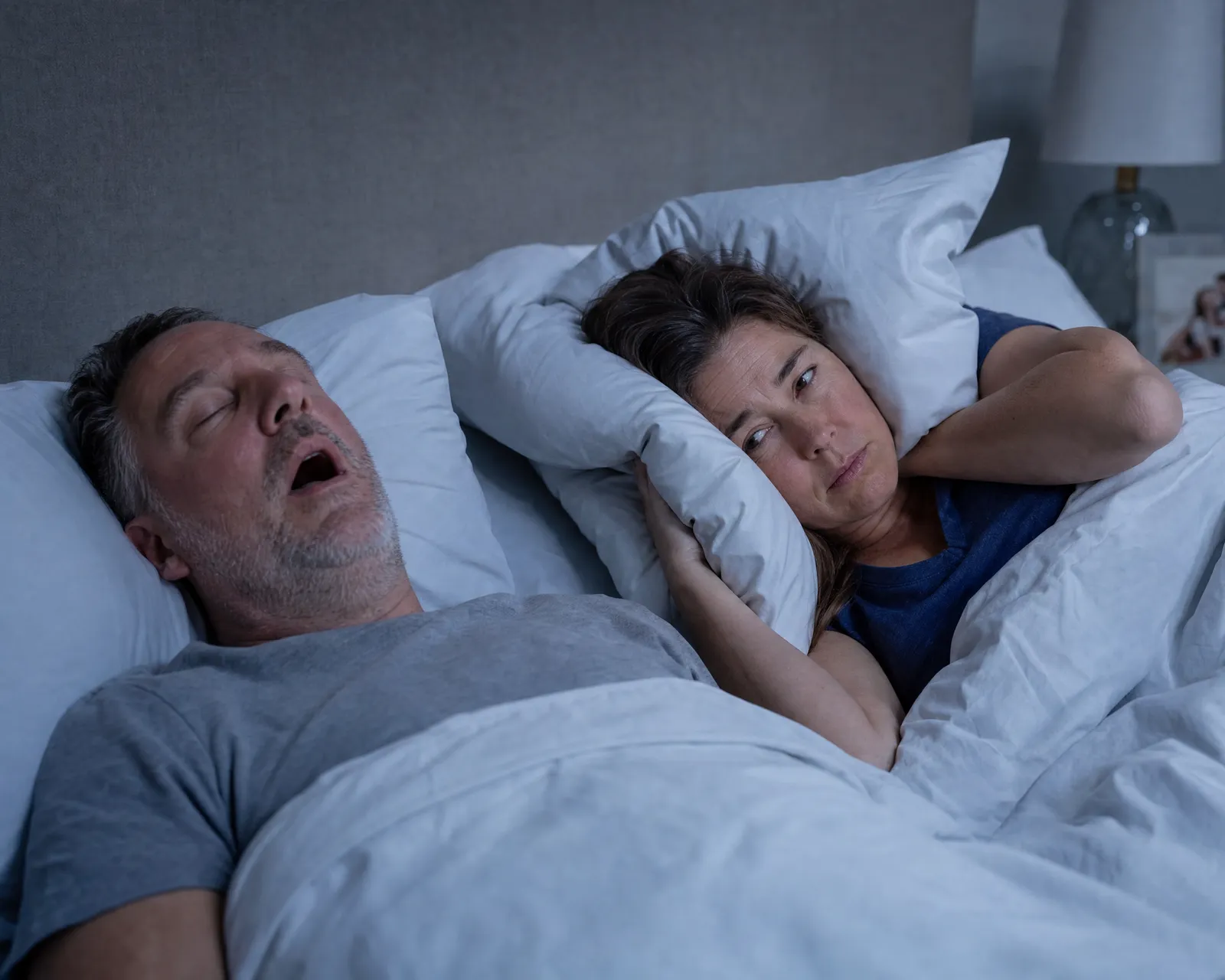 A man snoring in bed while his tired partner covers her ear with a pillow